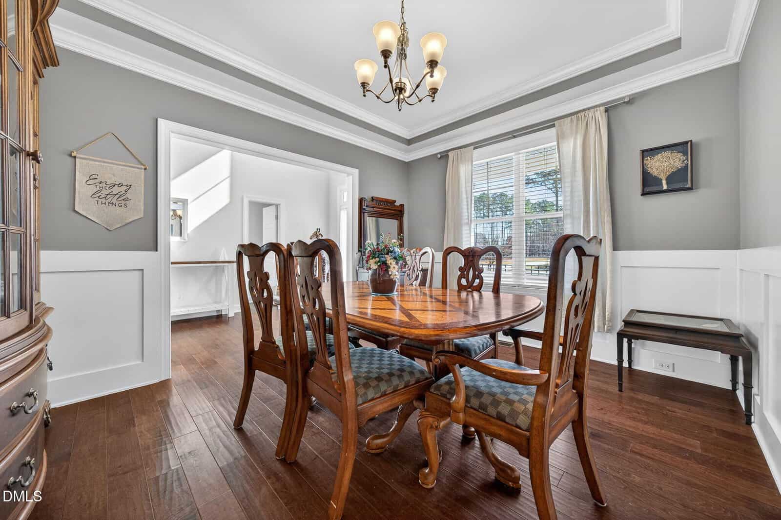A formal dining room with a wooden table and six chairs, hardwood floors, gray walls with white wainscoting, a large window with curtains, a chandelier, and a floral centerpiece on the table.