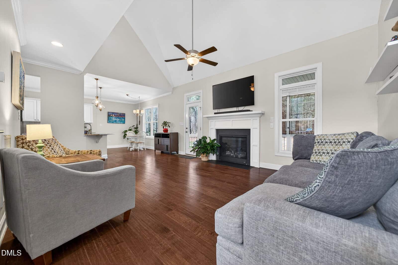 Spacious living room with wood flooring, gray sofa, armchair, fireplace with TV above, and large windows. Ceiling fans and recessed lighting provide illumination. Dining area visible in the background.