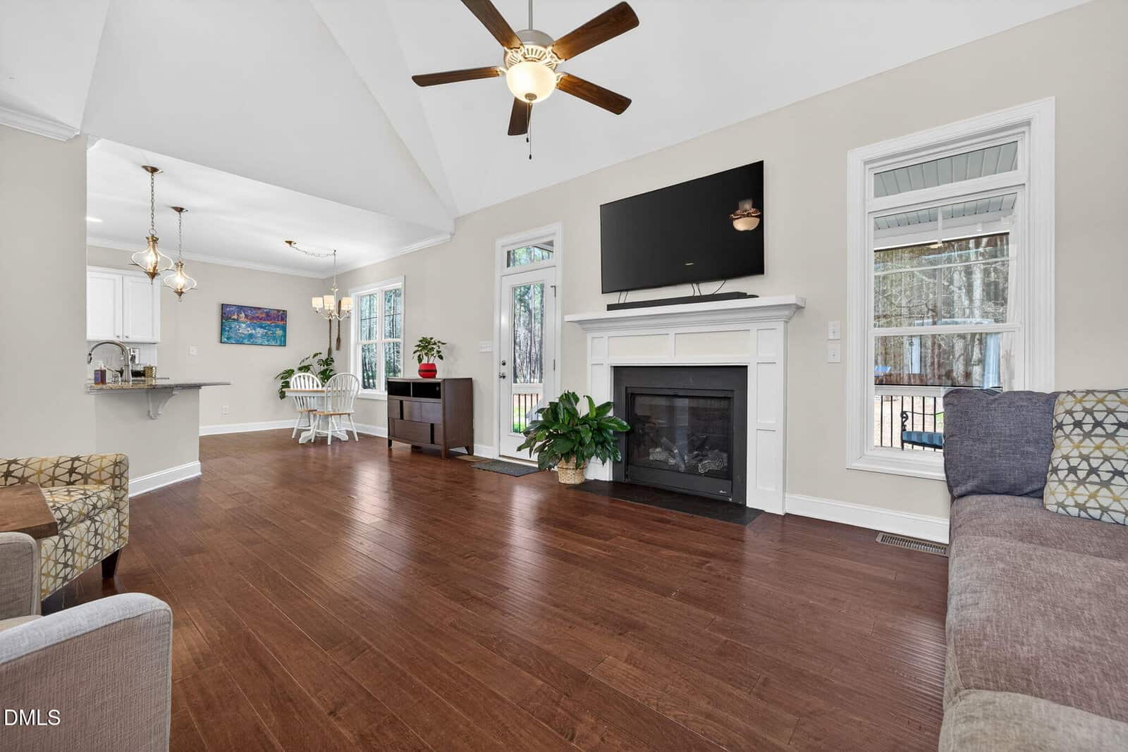 Bright living room with wood floors, a ceiling fan, fireplace with a TV above, windows, a sofa, plants, and an open dining area with a table and chairs leading into the kitchen.