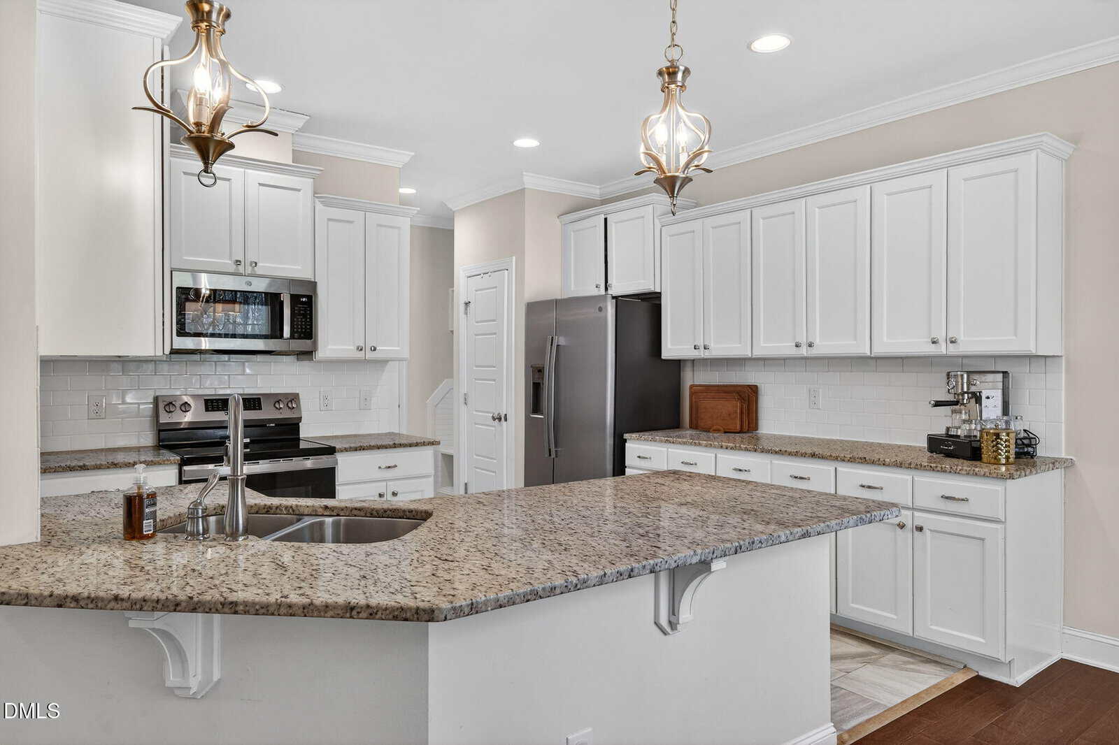 Modern kitchen with white cabinets, stainless steel appliances, granite countertops, and two pendant lights hanging above a large island with a built-in sink and faucet. Coffee maker and spice rack on the counter.