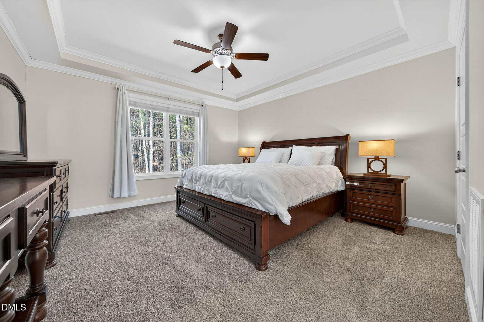 Spacious bedroom with beige walls, a large window with white curtains, a ceiling fan, and a wooden bed with white bedding flanked by matching nightstands and lamps. Carpeted floor and dresser also visible.