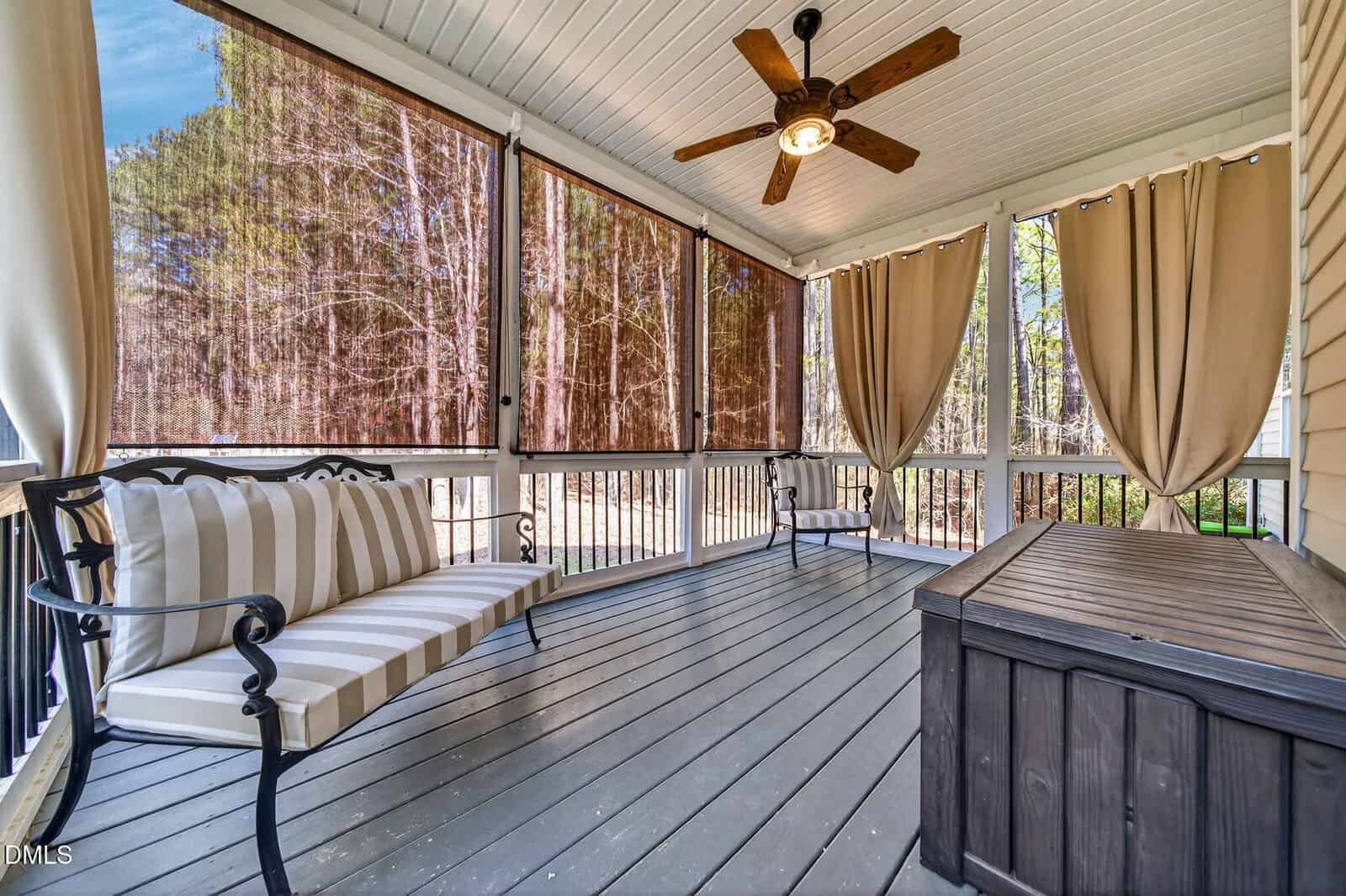 A covered porch with a ceiling fan, striped cushioned bench, black metal chair, wooden deck, storage chest, and large windows with rolled bamboo shades and beige curtains, overlooking a wooded area.