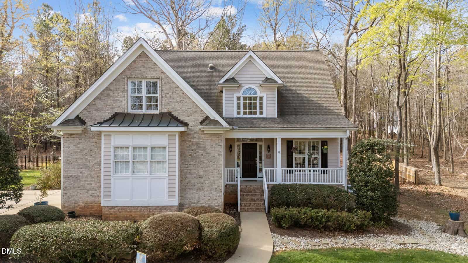 A two-story suburban house with brick and siding exterior, pitched roof, a covered front porch, white trim, and shrubbery in the front yard. The house is surrounded by trees and has a concrete walkway.