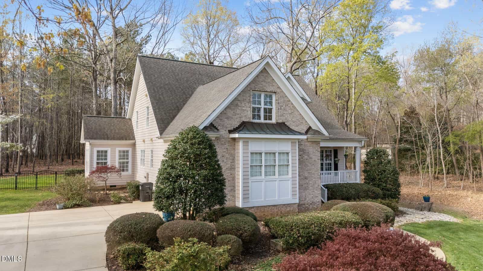 A two-story house with light brick and siding exterior, gable roof, front porch, and manicured shrubs. The house sits beside a wooded area and has a concrete driveway. The sky is partly cloudy.