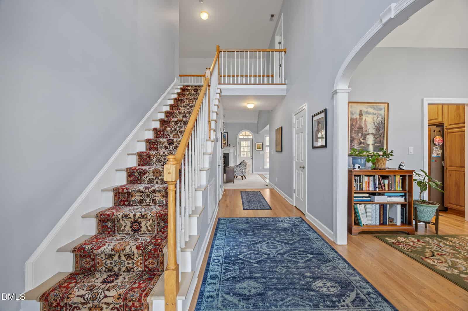 A hallway with light blue walls, wood flooring, and a patterned blue rug, featuring a staircase with a patterned carpet runner and wooden banister, leading to an open living area and a room with bookshelves and art.