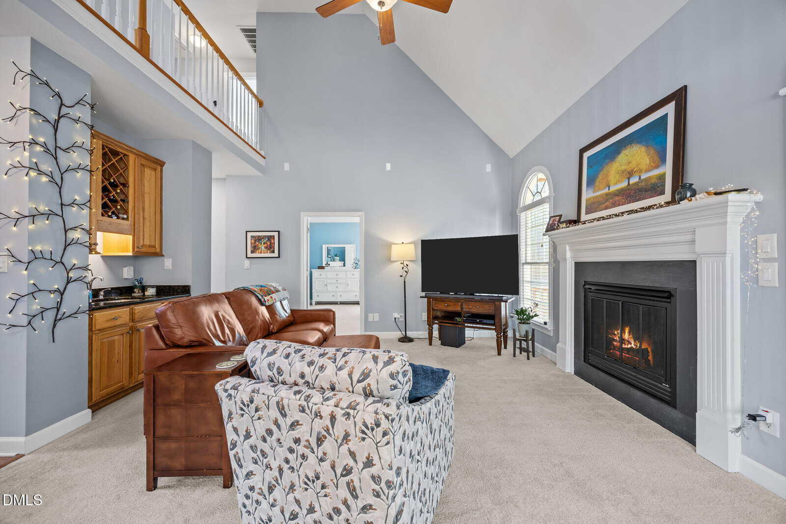 A cozy living room with a fireplace, leather sofa, patterned armchair, TV, and wet bar. Walls are light blue, ceiling is vaulted with a fan, and string lights decorate a tree-shaped structure in the corner.