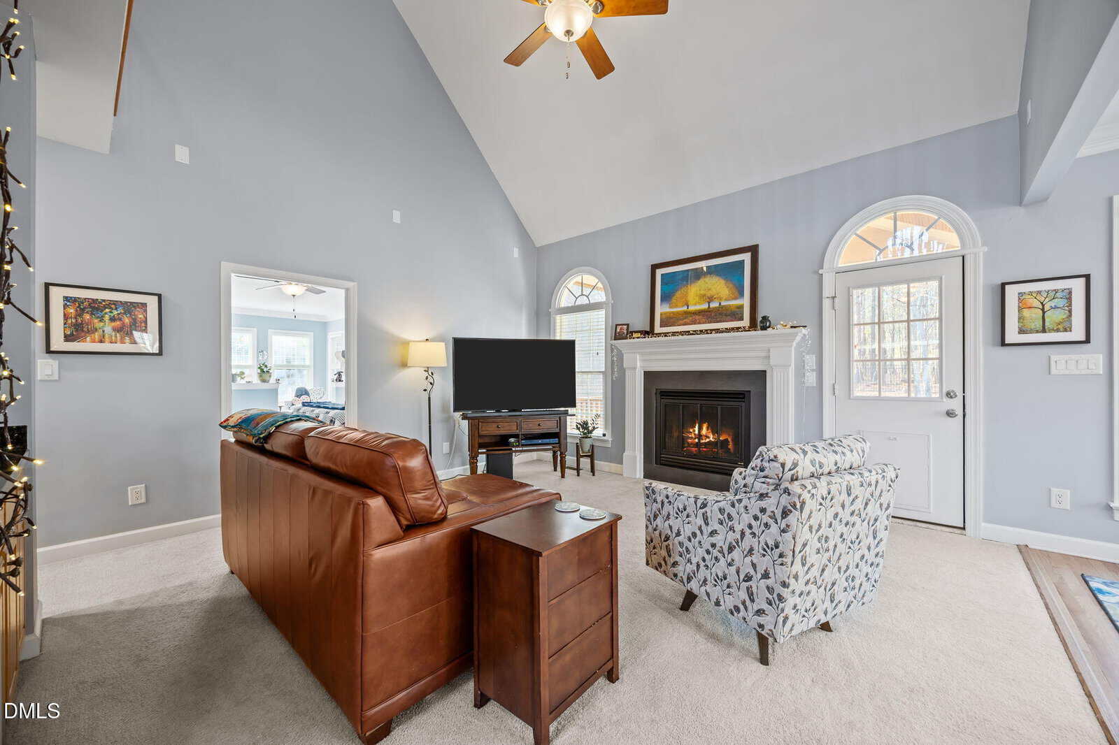Living room with high ceiling, brown leather sofa, patterned armchair, fireplace, TV, light walls, beige carpet, large windows, and framed artwork above the mantel. Ceiling fan and natural light brighten the space.