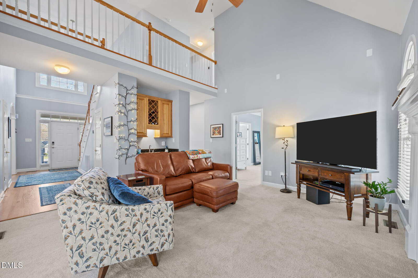Spacious living room with tan carpet, leather sectional, floral armchair, large TV, and bright natural light. Overlooks open hallway upstairs and entryway with white front door.