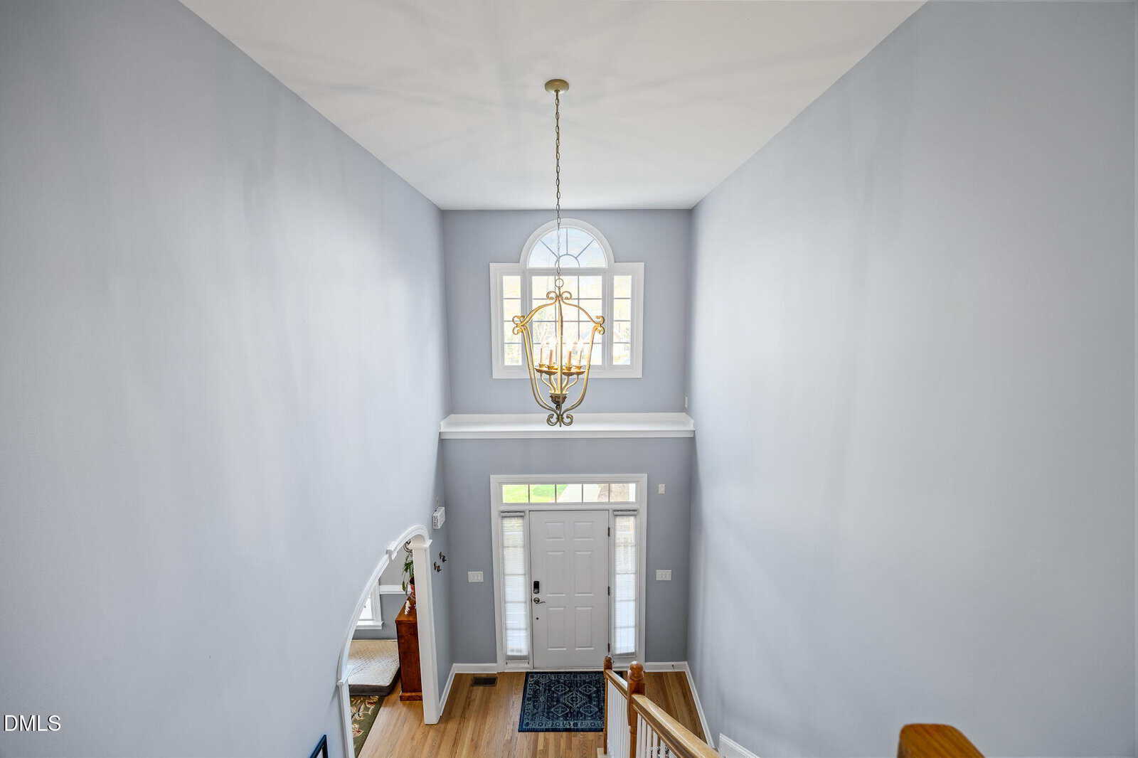 A view from the top of a staircase shows a foyer with light gray walls, a white front door, a large window above the door, and a hanging chandelier. A wooden floor and partial view of adjacent rooms are visible.