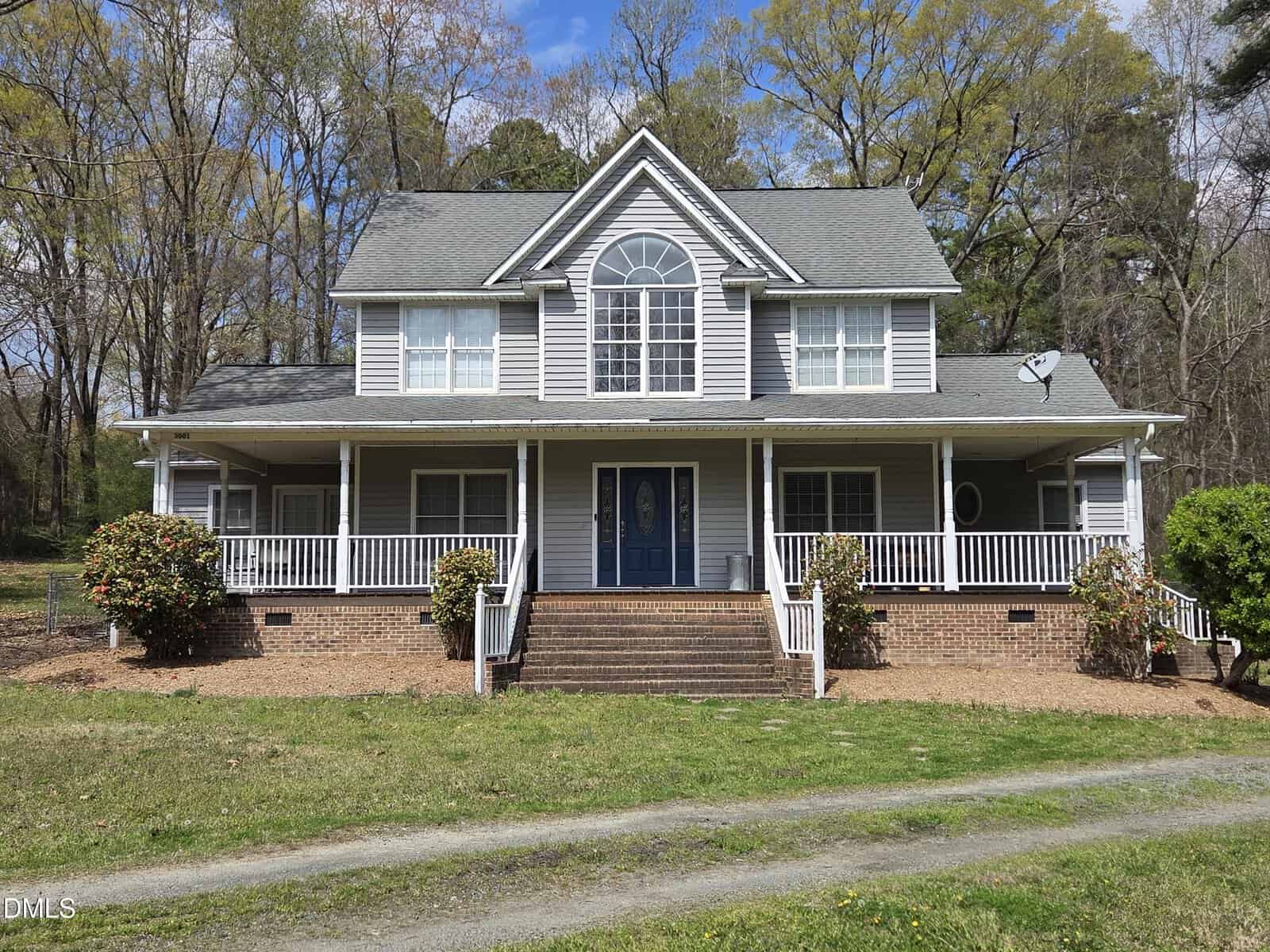 Two-story gray house with white trim, large front porch, and gabled roof. Double front doors with an arched window above. Surrounded by bushes, trees, and a gravel driveway.