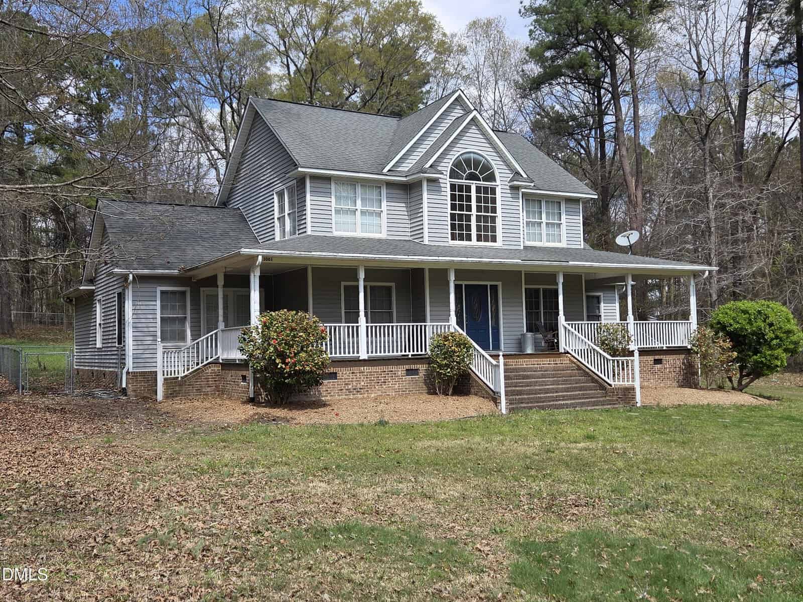 A two-story house with gray siding, white trim, a covered wraparound porch, and a gable roof, set on a grassy yard with bushes and surrounded by tall trees.