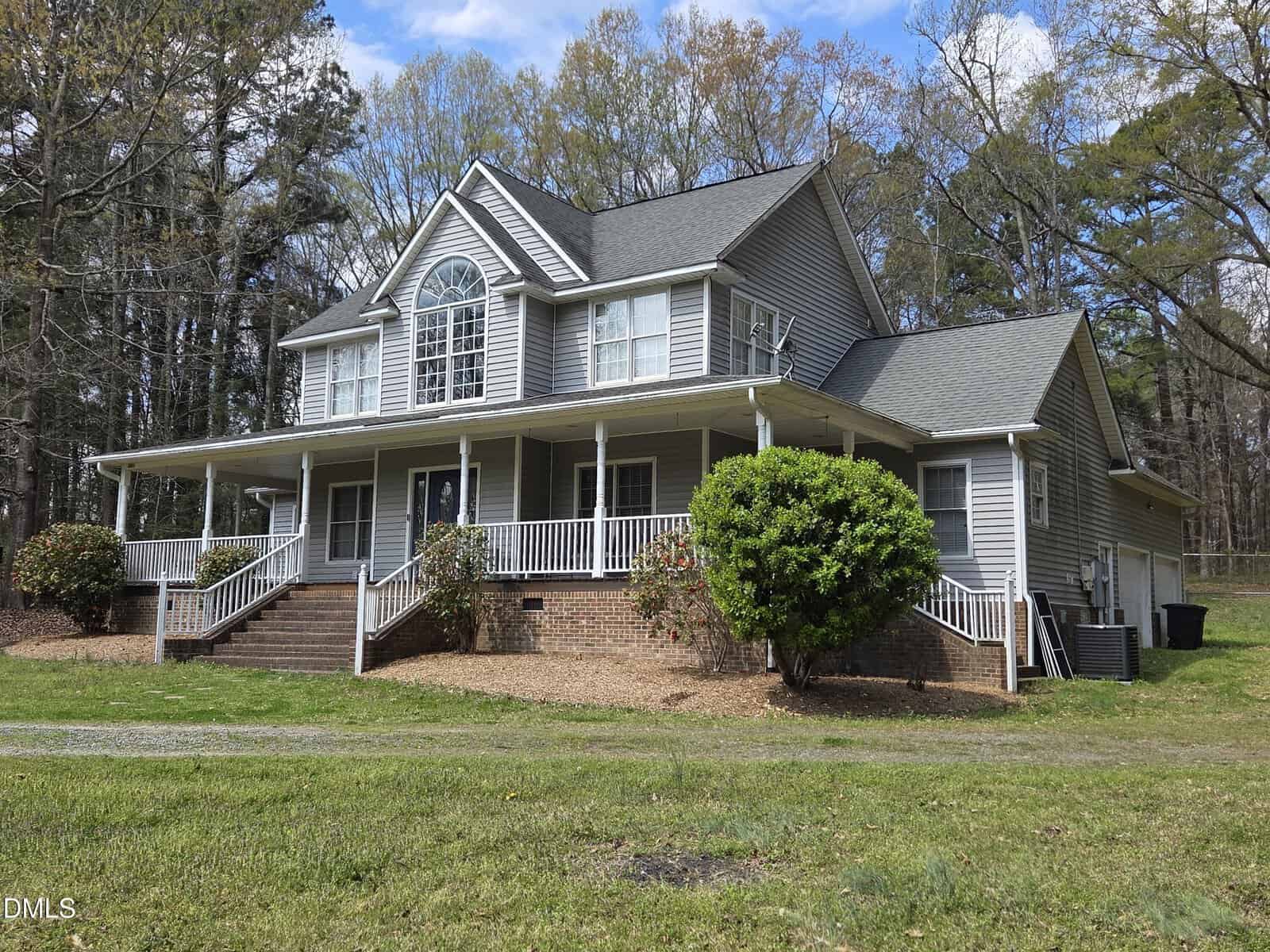 Two-story gray house with white trim and a large front porch, set on a grassy lot with trees in the background. Stairs lead to the porch, and there are shrubs and mulch around the house.