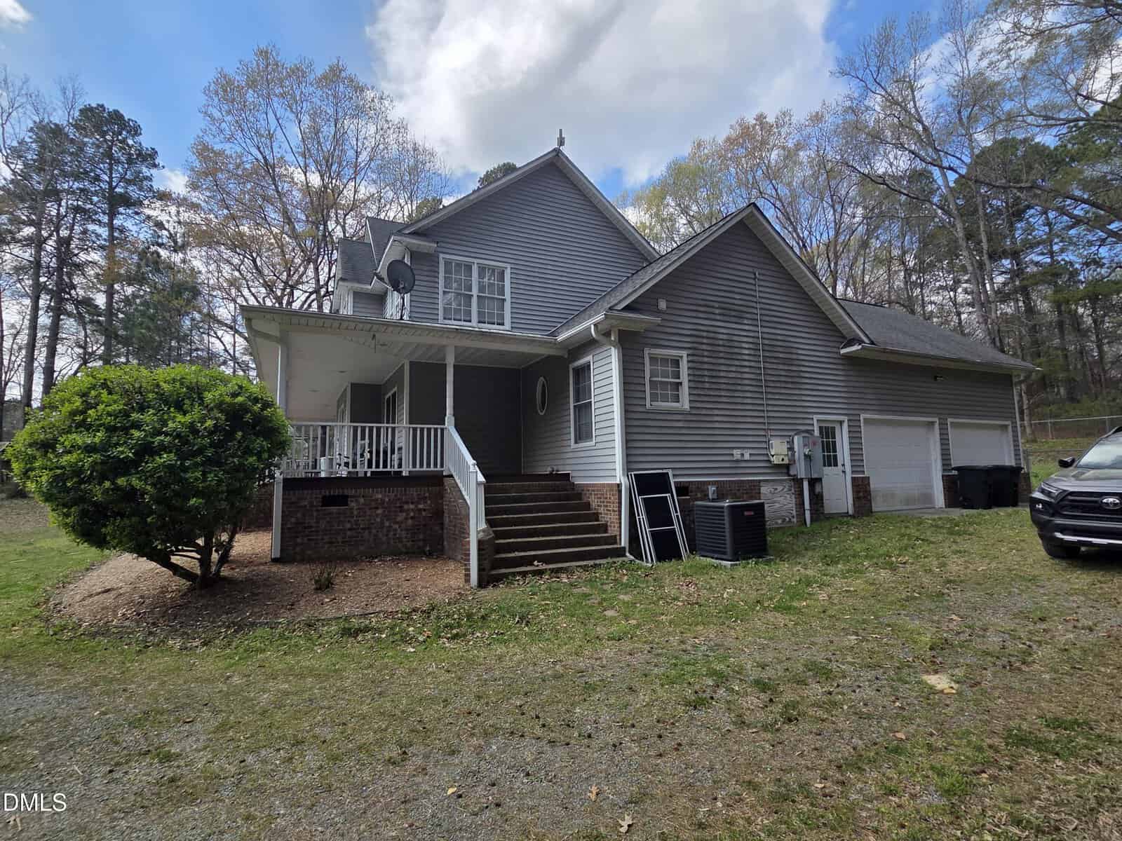 Two-story gray house with white trim, small covered porch, brick steps, two garage doors, two HVAC units, satellite dish, and a black car parked to the right on a gravel driveway. Trees and cloudy sky in the background.