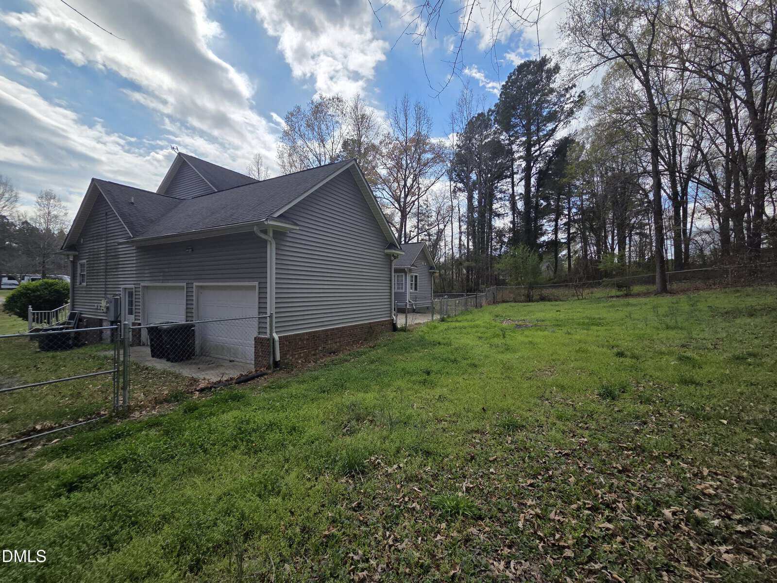 A gray house with a fenced yard, double garage, and a grassy area bordered by trees under a partly cloudy sky.