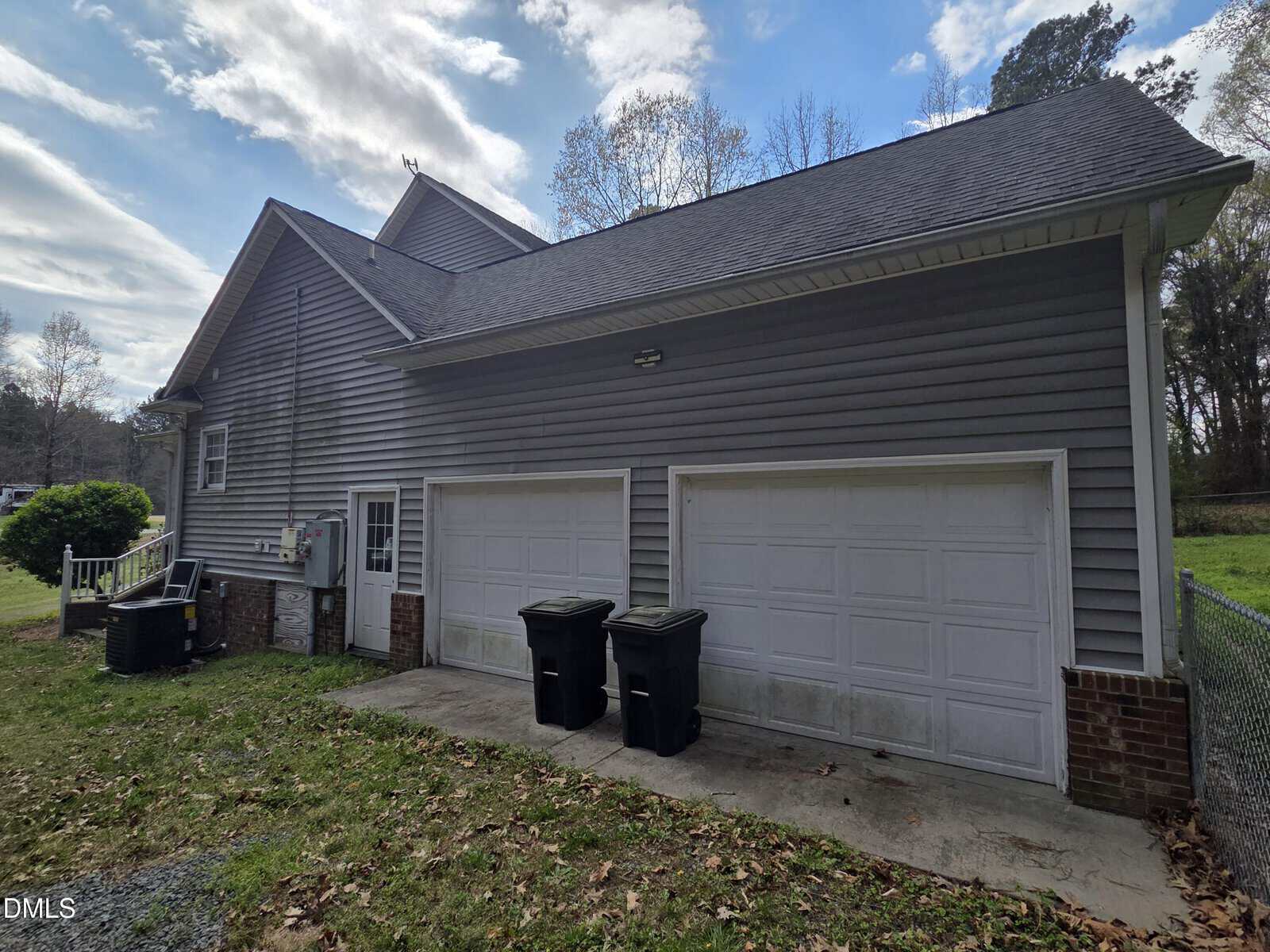 A gray house with a double garage, two white garage doors, two black trash bins, and a fenced yard. The roof is dark gray and the siding is horizontal vinyl. The sky is partly cloudy with trees in the background.