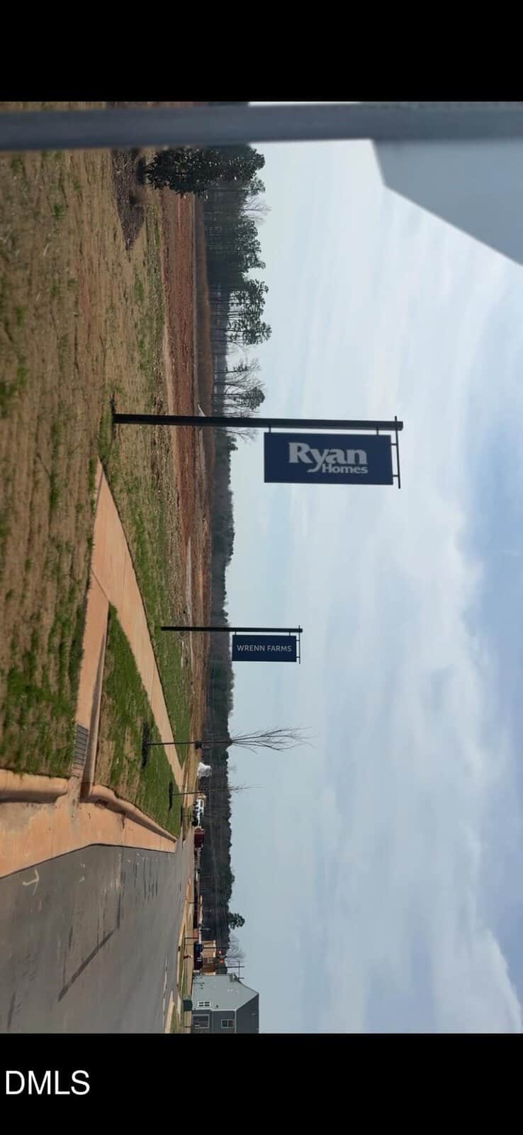 A paved road with sidewalks runs through a new housing development. Signs along the road display "Ryan Homes" and "Wrenn Farms." The area has grass, bare land, and a few houses in the distance.