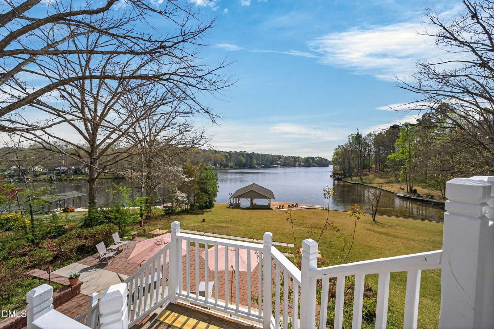 View from white-railed stairs overlooking a lawn, pavilion, and calm lake surrounded by trees under a blue sky with clouds. Lounge chairs and brick patio area visible in the foreground.