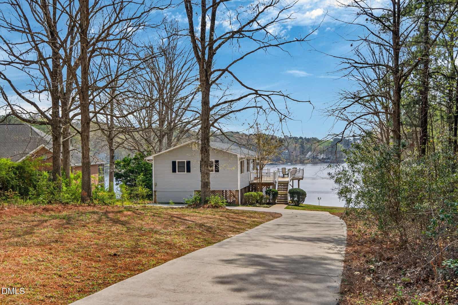 A concrete driveway leads to a white house with a deck overlooking a lake. Leafless trees line the yard, and docks and other houses are visible across the water under a partly cloudy sky.