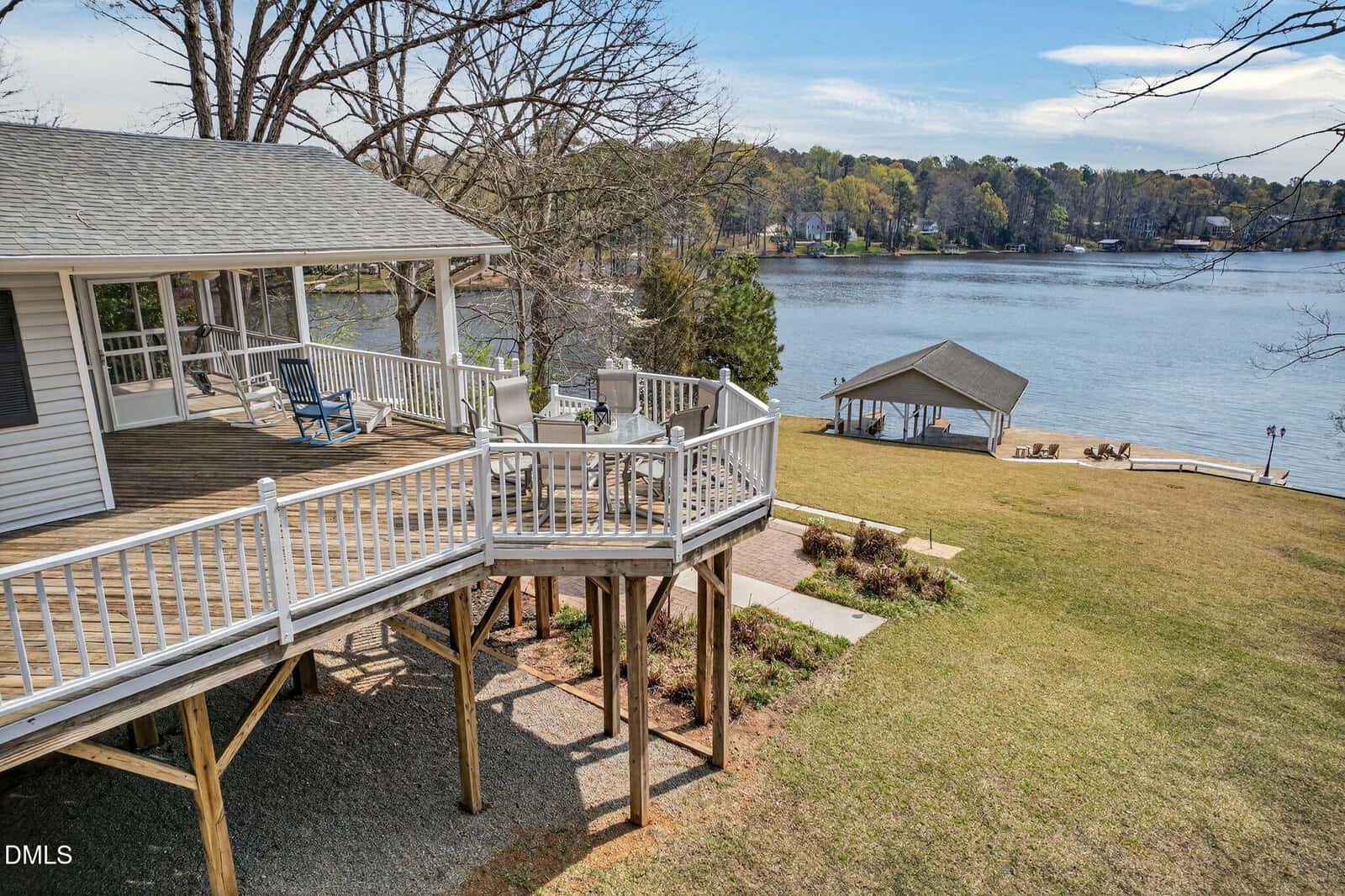 Wooden deck with white railings attached to a house overlooks a grassy yard leading to a lake. A covered dock is visible on the water, and there are chairs and outdoor furniture on the deck. Trees and neighboring houses in the background.