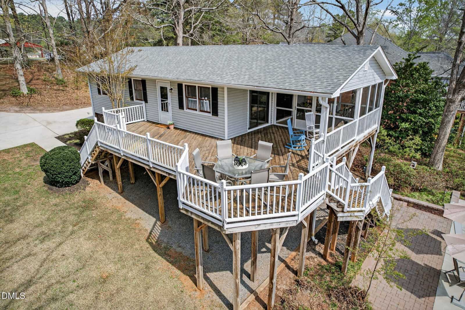 A single-story white house with a large wooden deck on stilts, featuring railings, outdoor furniture, and steps leading to a brick patio. The house is surrounded by trees and a landscaped yard.