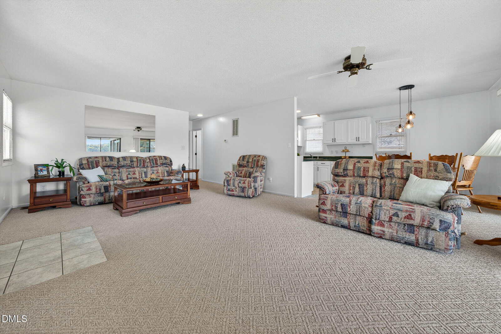 Spacious living room with patterned sofas, recliner, coffee table, and neutral carpet. Open view to dining area and kitchen with white cabinets. Ceiling fans and large windows provide light.