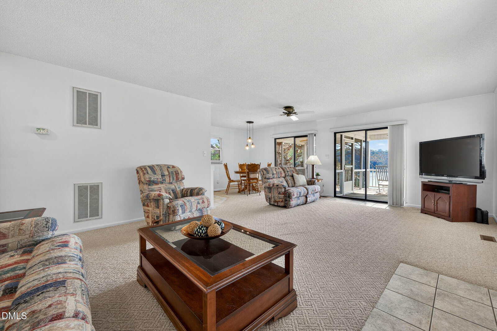 Spacious living room with patterned sofas, coffee table, TV, and sliding glass doors leading to a balcony. Dining area and ceiling fan are visible in the background. Neutral colors and carpeted floors throughout.