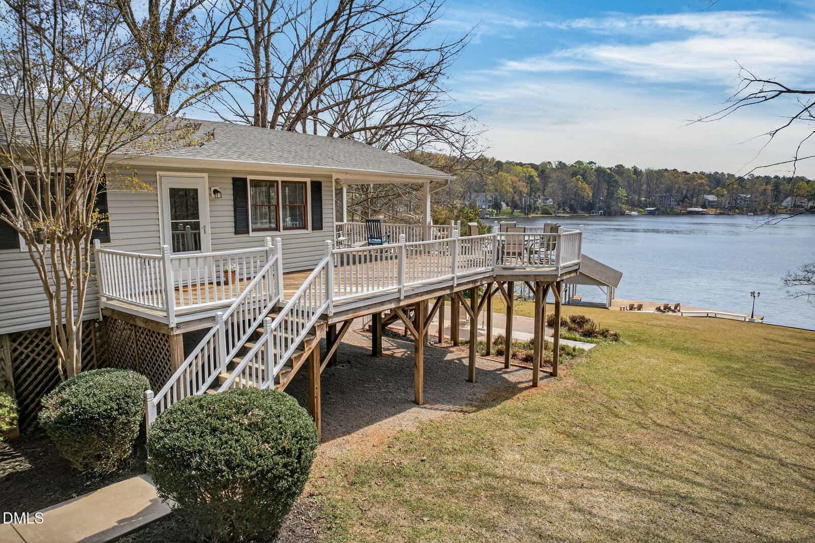 A raised house with a large wooden deck and stairs overlooks a lake. The yard is sloped with trimmed bushes near the house. Trees border the property, and the lake and distant shoreline are visible under a partly cloudy sky.