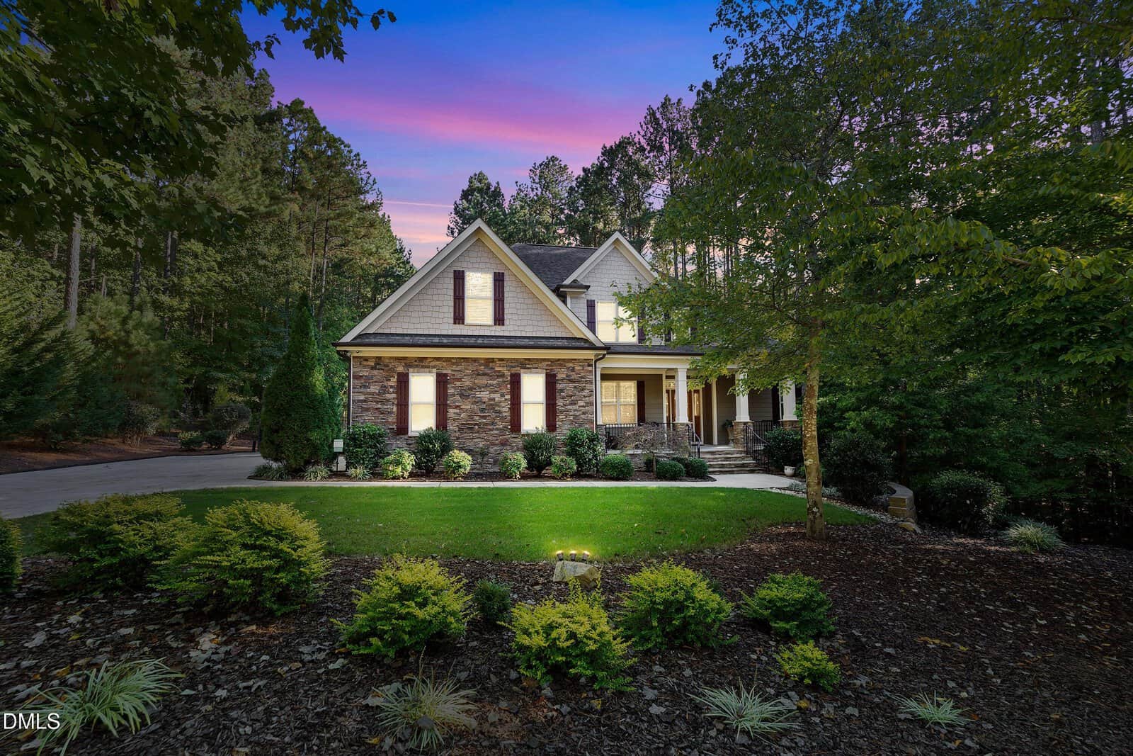A two-story house with stone and siding exterior, surrounded by trees and landscaped bushes, is beautifully showcased at sunset with a purple and pink sky at 1202 New Grissom Way in Wake Forest.