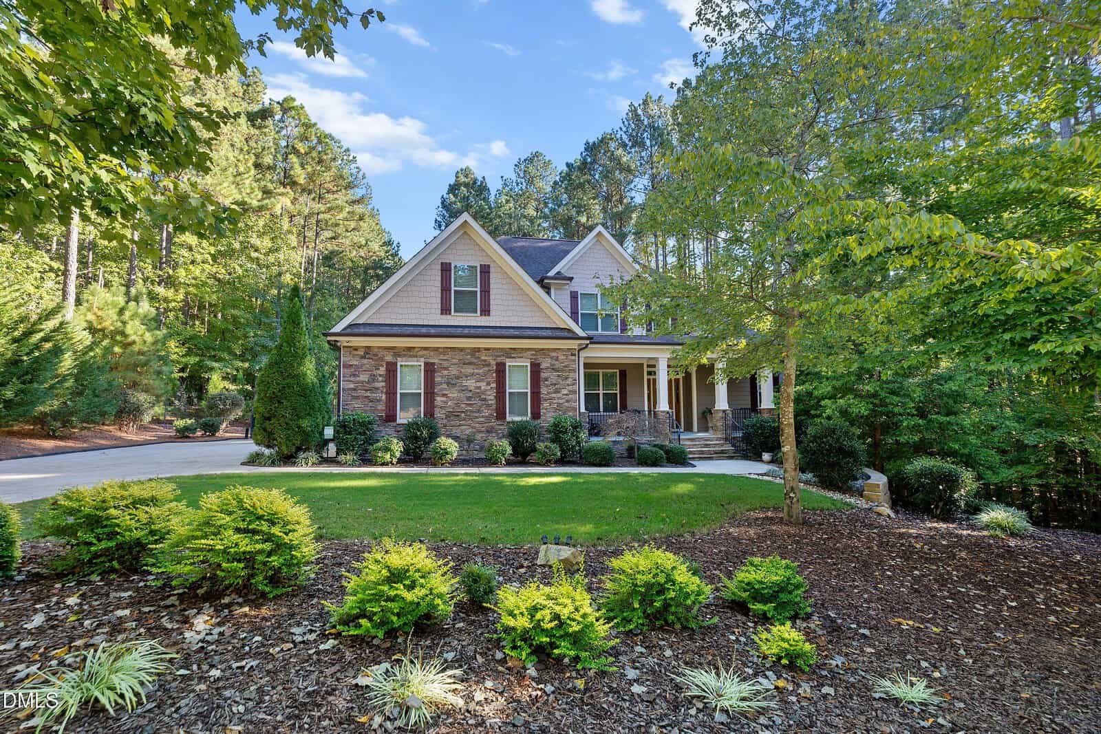 Two-story house with brick and siding exterior, surrounded by trees and landscaped front yard with shrubs and grass. Concrete driveway curves to the left. Blue sky with scattered clouds above.