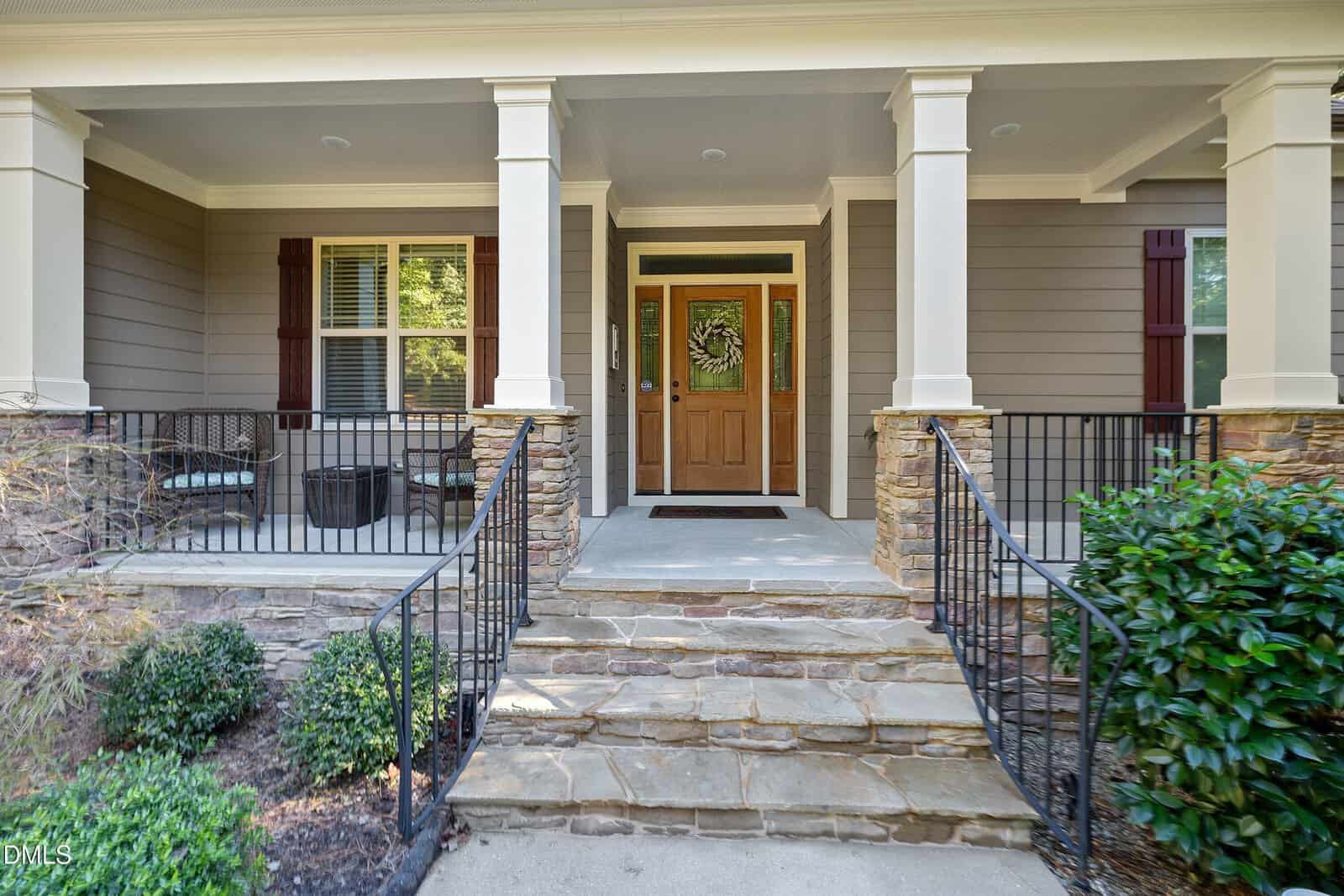 Stone steps lead to a covered front porch with a wooden door, side windows, decorative columns, and a seating area. Bushes and plants line the walkway.