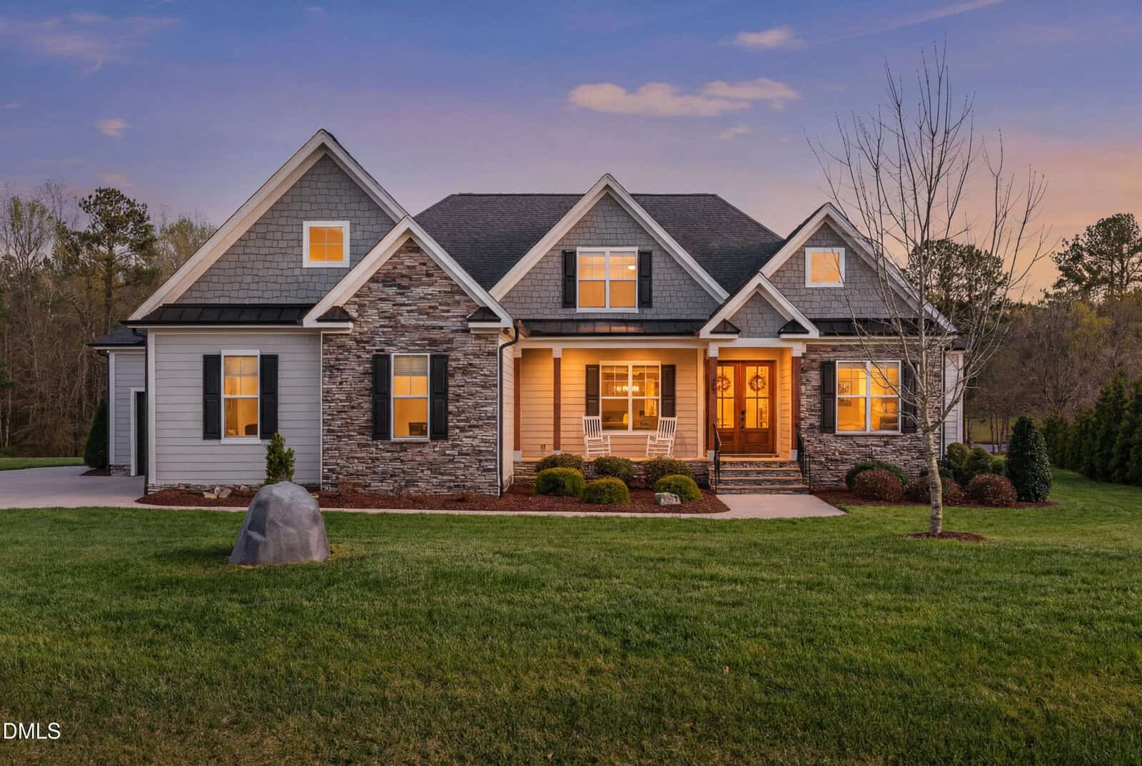 A modern suburban house at 90 Old Spur Road, Youngsville, with stone and siding exterior, large glowing windows, a front porch with rocking chairs, well-kept lawn at dusk, plus a leafless tree and landscaping in the front yard.