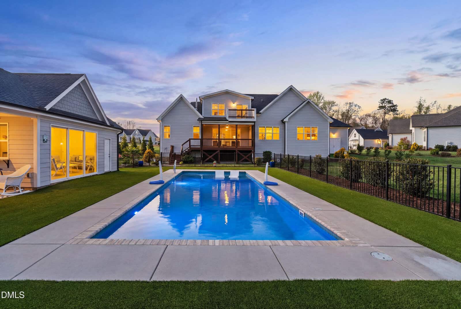 A modern two-story house with large windows and a spacious backyard featuring a rectangular swimming pool, surrounded by manicured lawn and a patio area, at sunset.