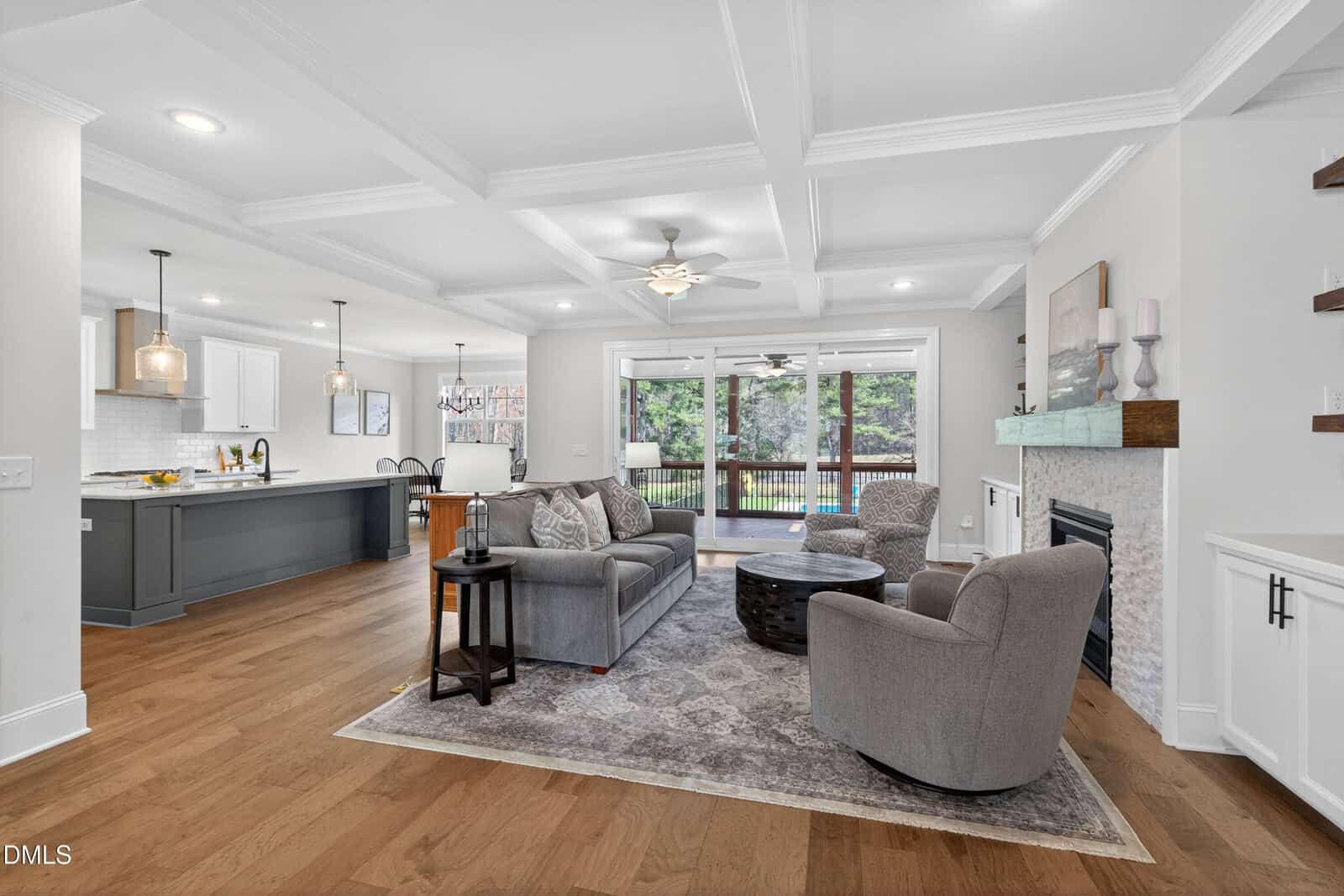 Open-concept living room with gray sofas, a round coffee table, fireplace, and large windows leading to a deck. The space connects to a modern kitchen with pendant lights and white cabinetry.