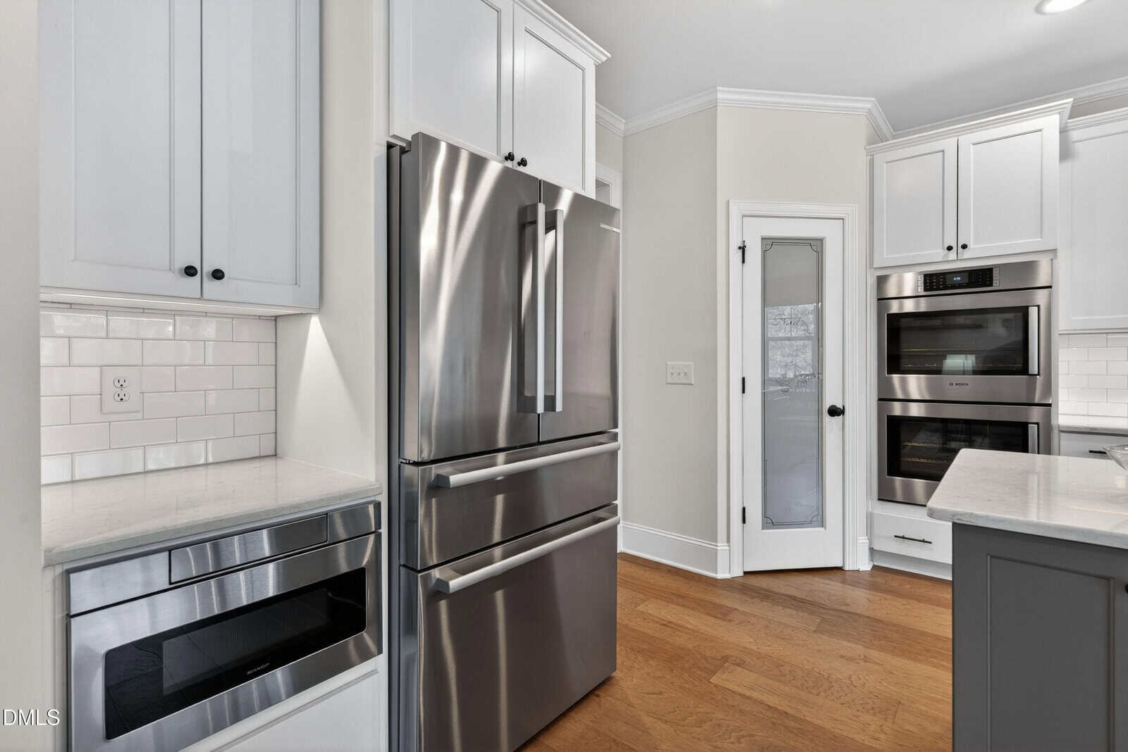 Modern kitchen with stainless steel refrigerator, built-in oven, microwave, white cabinets, subway tile backsplash, and a pantry door with frosted glass, all set on a wooden floor.