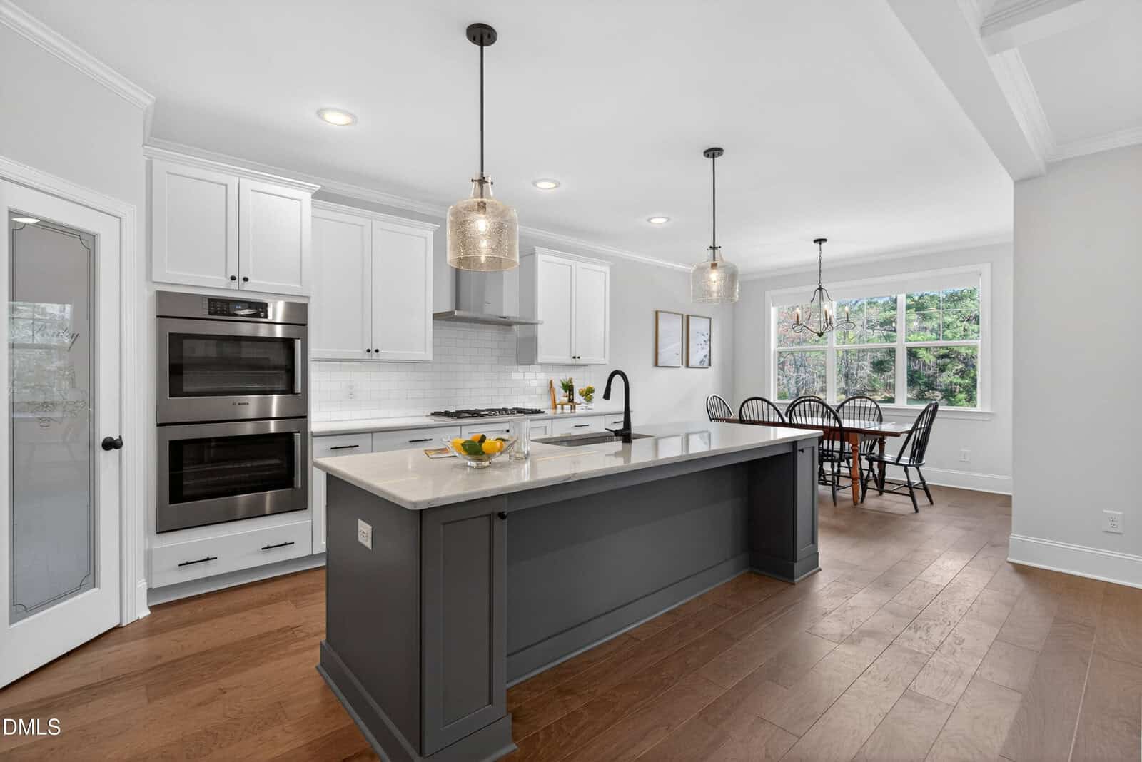 Modern kitchen with white cabinets, a gray island, built-in double ovens, pendant lights, and a dining area by large windows overlooking greenery. Wood flooring throughout.