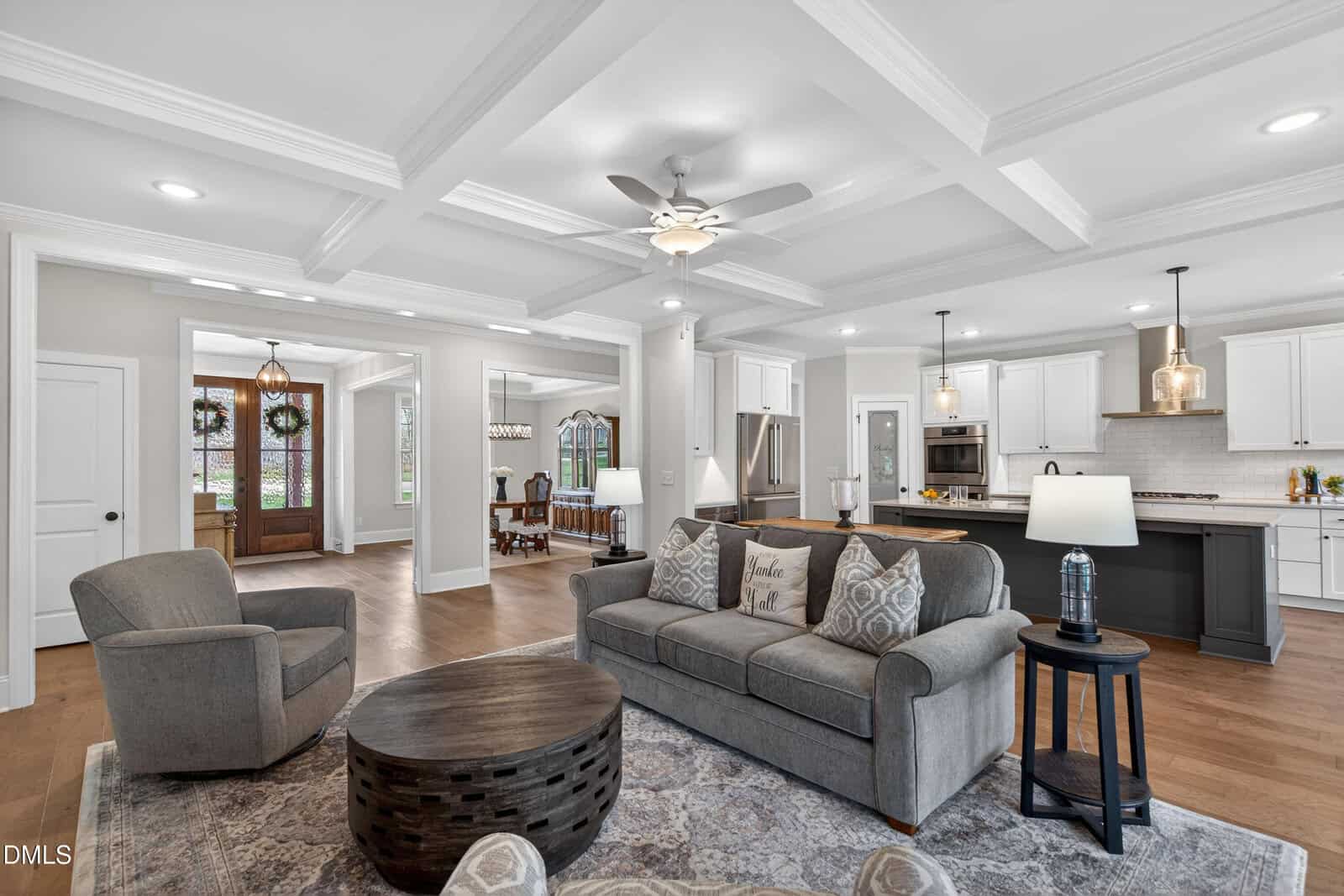 A spacious, modern living room with a gray couch, armchair, round coffee table, and open layout leading to a bright kitchen with white cabinets and an adjacent dining area. The room features coffered ceilings and wood flooring.