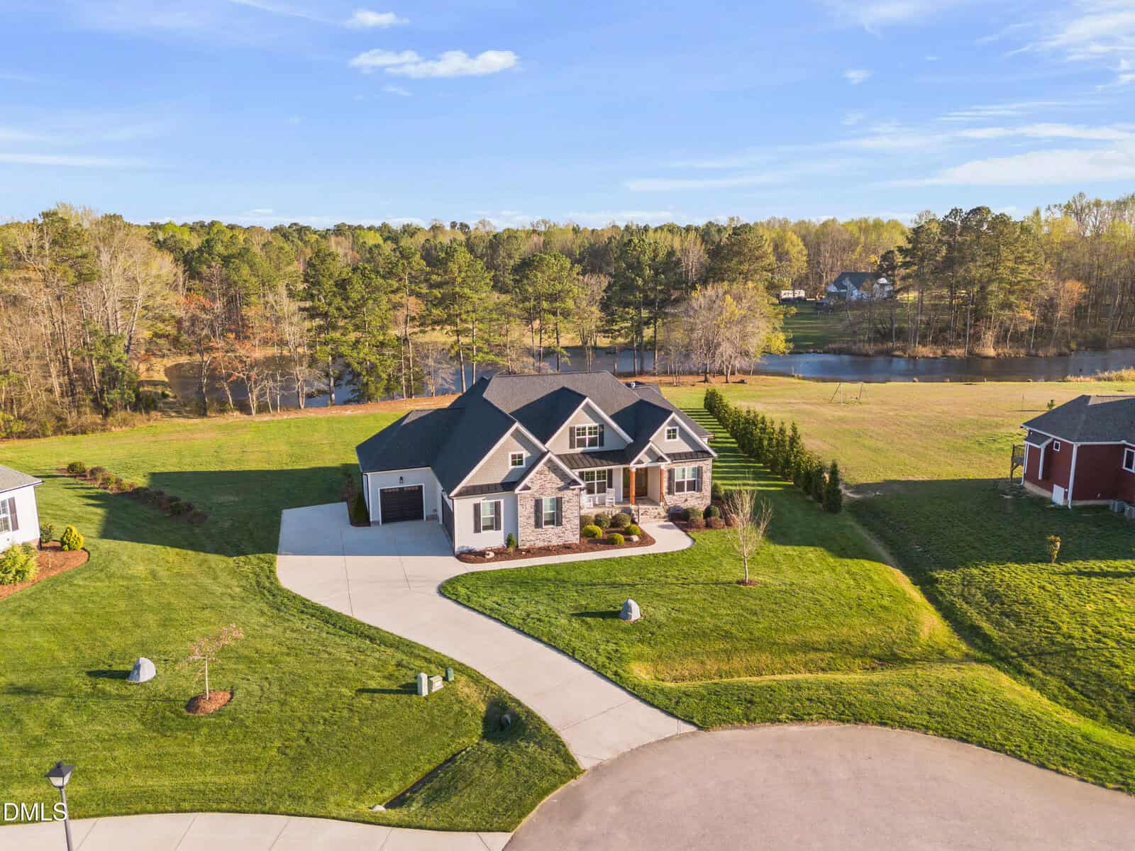 A large house with a dark roof and light exterior sits on a spacious, well-manicured lawn near a curved driveway, with a pond, trees, and neighboring homes in the background under a clear sky.