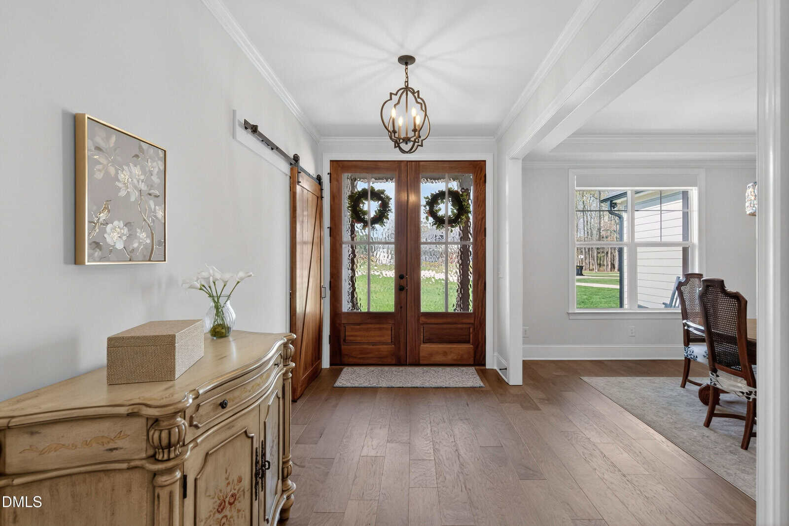 Bright entryway with wooden double doors decorated with wreaths, chandelier overhead, wooden floors, a light-colored cabinet with flowers and a box, and an adjacent room visible with a dining table and chairs.