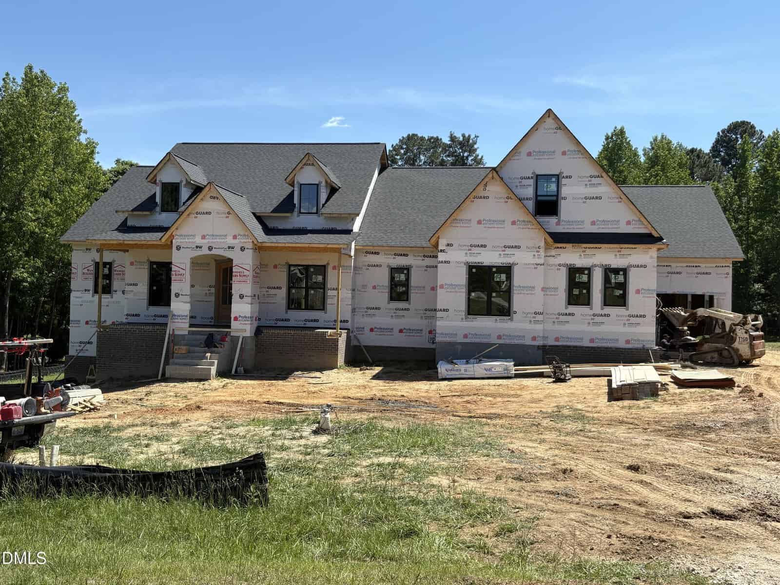 A large house under construction with exposed framing and house wrap, multiple gabled roofs, and construction materials scattered around the dirt yard. Trees and greenery are visible in the background.