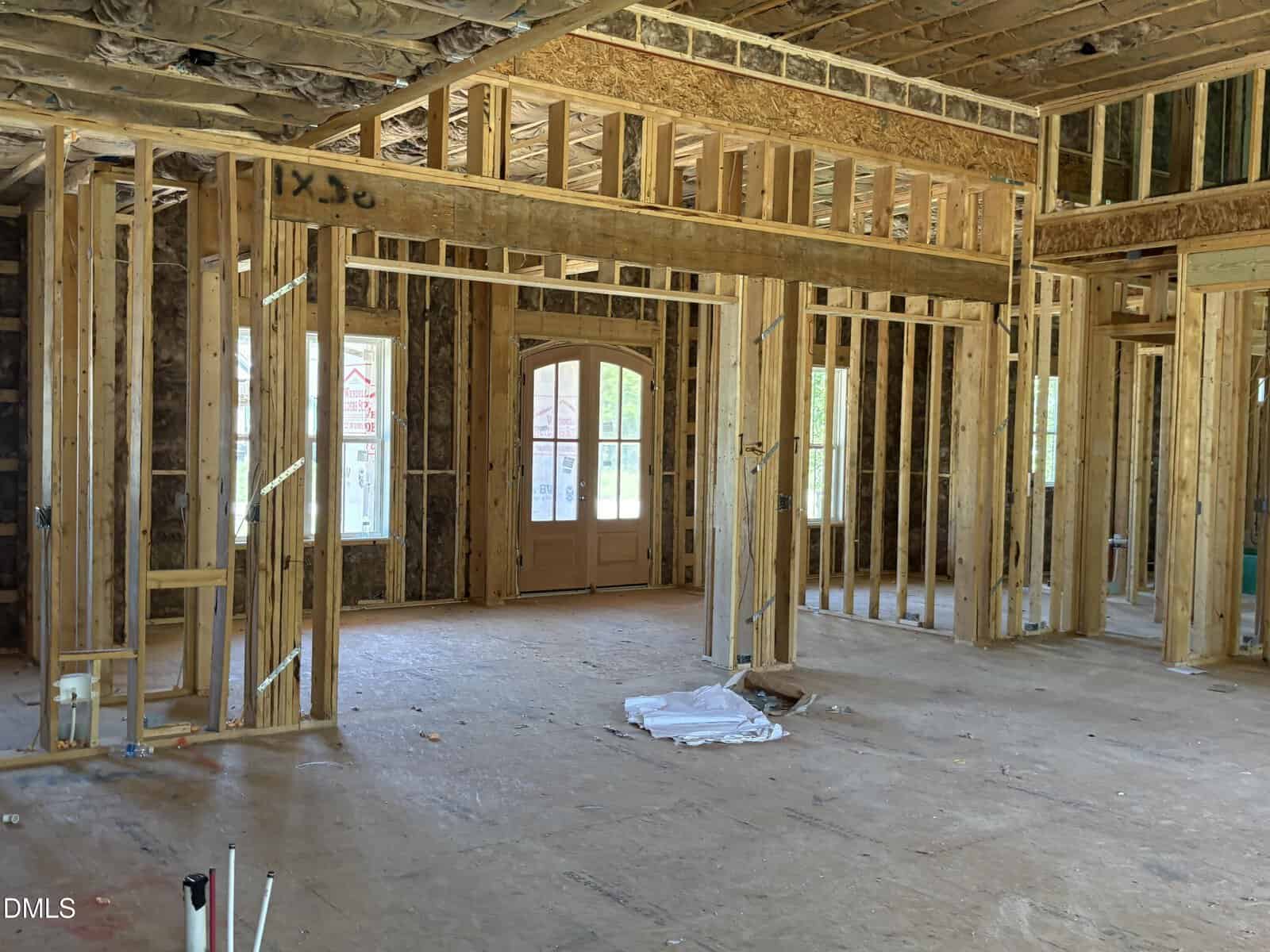 Interior of a house under construction showing exposed wooden framing, unfinished walls, large windows, and a double front door. The floor is bare, and building materials are scattered around.