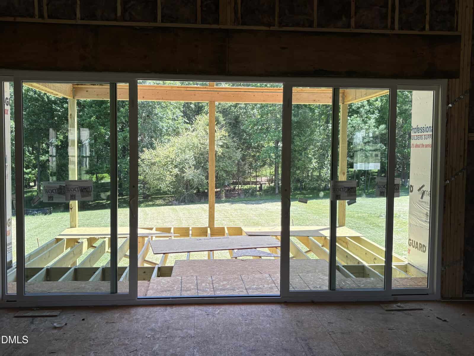 View through large glass sliding doors shows a partially built wooden deck and a green, wooded backyard under a clear, sunny sky. Construction materials and tools are visible on the deck.