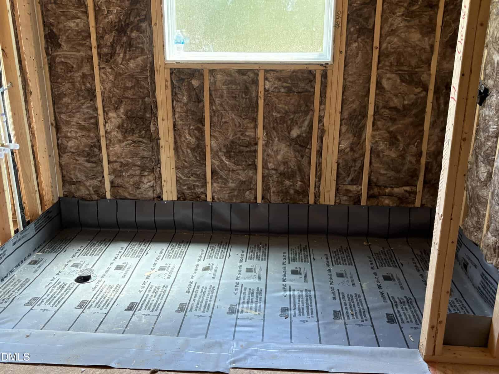 A shower under construction with exposed wall studs, insulation, and a newly installed waterproof liner on the floor. A window is centered in the wall above the shower area.
