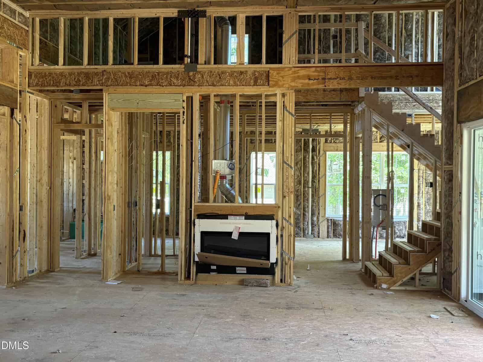 Interior view of a house under construction, showing exposed wooden framing, unfinished walls, a fireplace installation, and a staircase leading to the second floor. Sunlight comes through large windows.