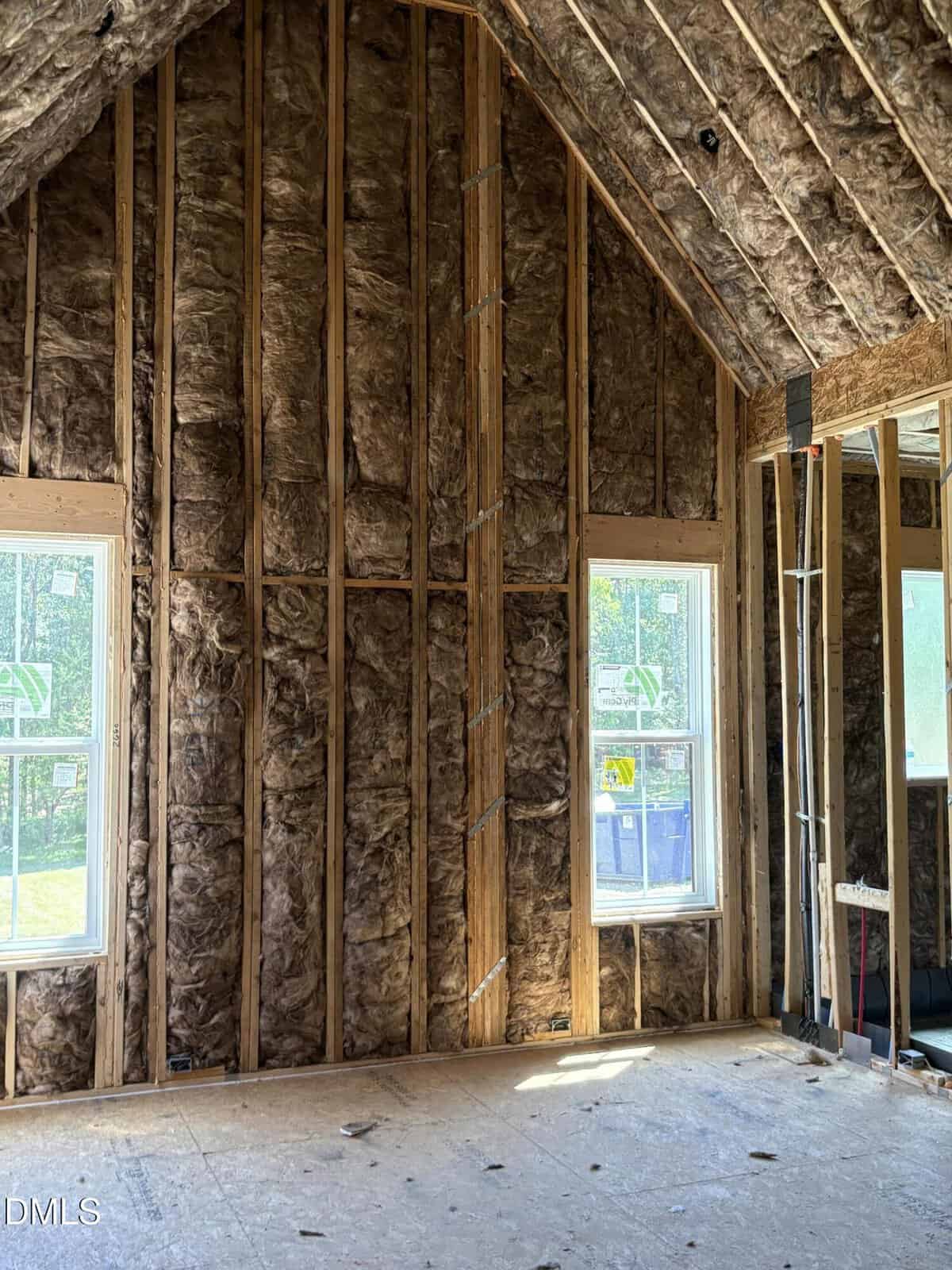 Interior of a house under construction showing exposed walls and ceiling lined with insulation material, two windows letting in natural light, and an unfinished wooden floor.