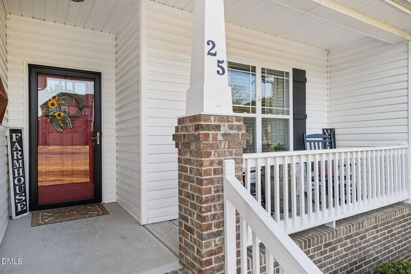 A front porch with white siding and a brick column, featuring a red front door with a sunflower wreath, a black "Farmhouse" sign, and a rocking chair beside a window with black shutters. The number 25 is on the post.