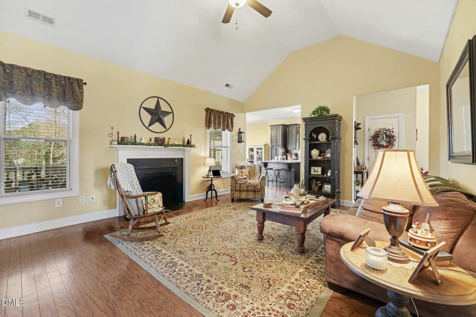 A living room with yellow walls, a vaulted ceiling, wooden floors, a fireplace with a star decoration above it, a brown sofa, a rocking chair, a rug, and a coffee table. The kitchen is visible through an open doorway.