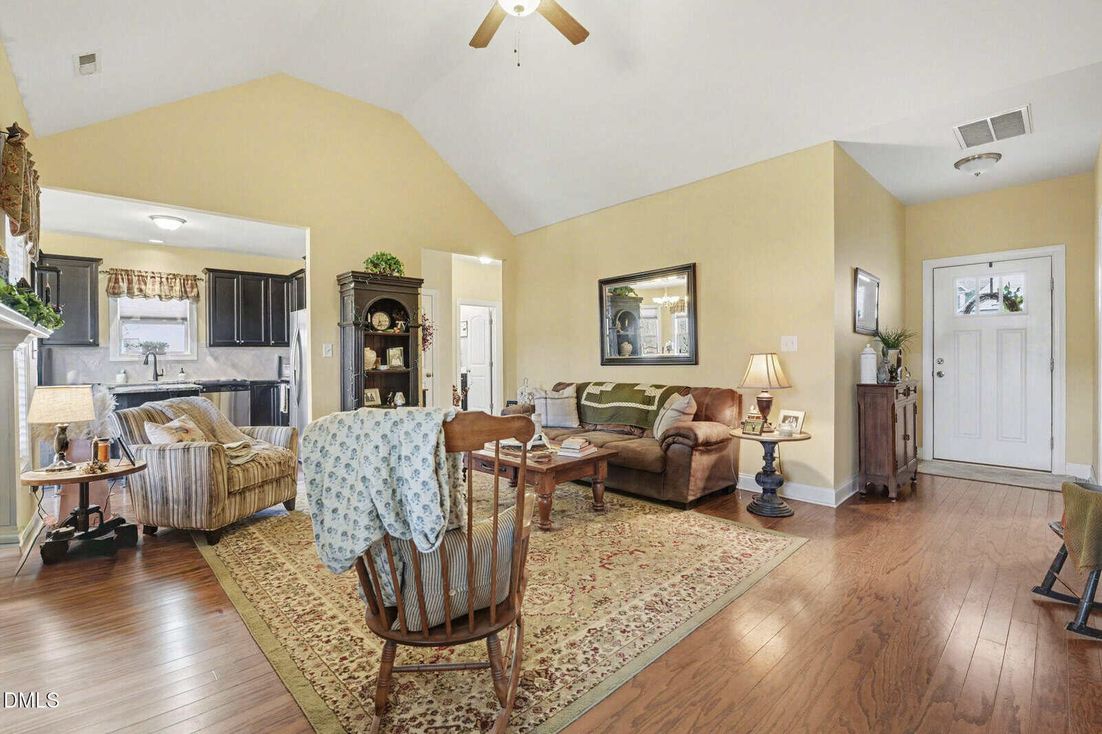 A living room with yellow walls, hardwood floors, and a high ceiling. It features a sofa, armchair, coffee table, and area rug. The front door, a small cabinet, and a view into a kitchen are visible.