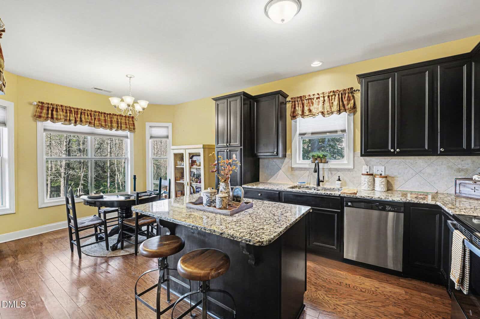Bright kitchen with yellow walls, dark cabinets, granite countertops, a center island with stools, stainless steel appliances, and a dining area by large windows with wooded views. Dining table seats four.