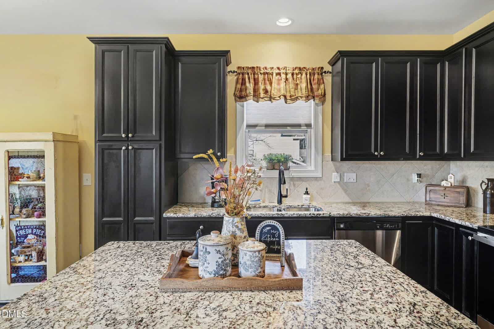 Modern kitchen with dark cabinets, granite countertops, and a window with a plaid curtain. A tray with jars and flowers sits on the island. A cabinet with groceries is on the left side.