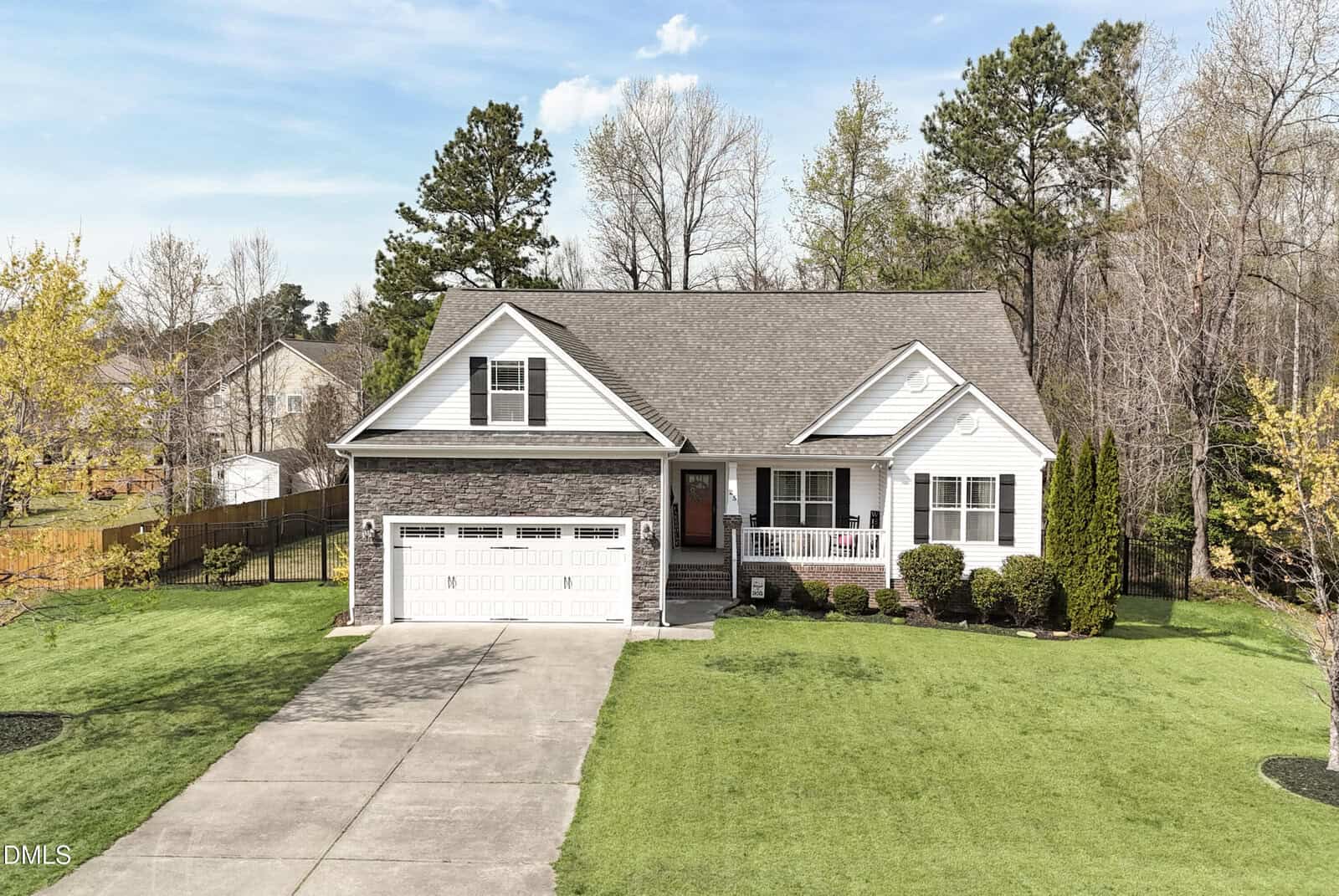 A single-story suburban house with a gray shingle roof, white siding, stone accents, and an attached two-car garage. The front yard has neatly trimmed grass, shrubs, and a concrete driveway. Trees border the property.